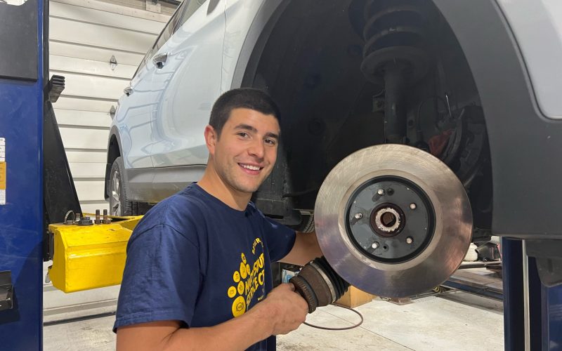 Car technician smiling while working on brake assembly, featuring a vehicle on a lift and brake rotor in focus, emphasizing automotive repair services related to brake maintenance.