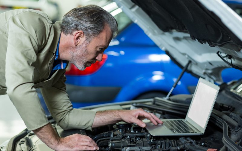 Mechanic using laptop while servicing car engine in repair garage