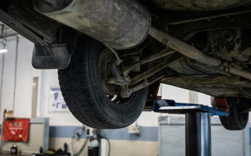 Underside view of a vehicle showing the control arms, tire, and suspension system in an automotive repair shop, emphasizing vehicle maintenance and control arm types.